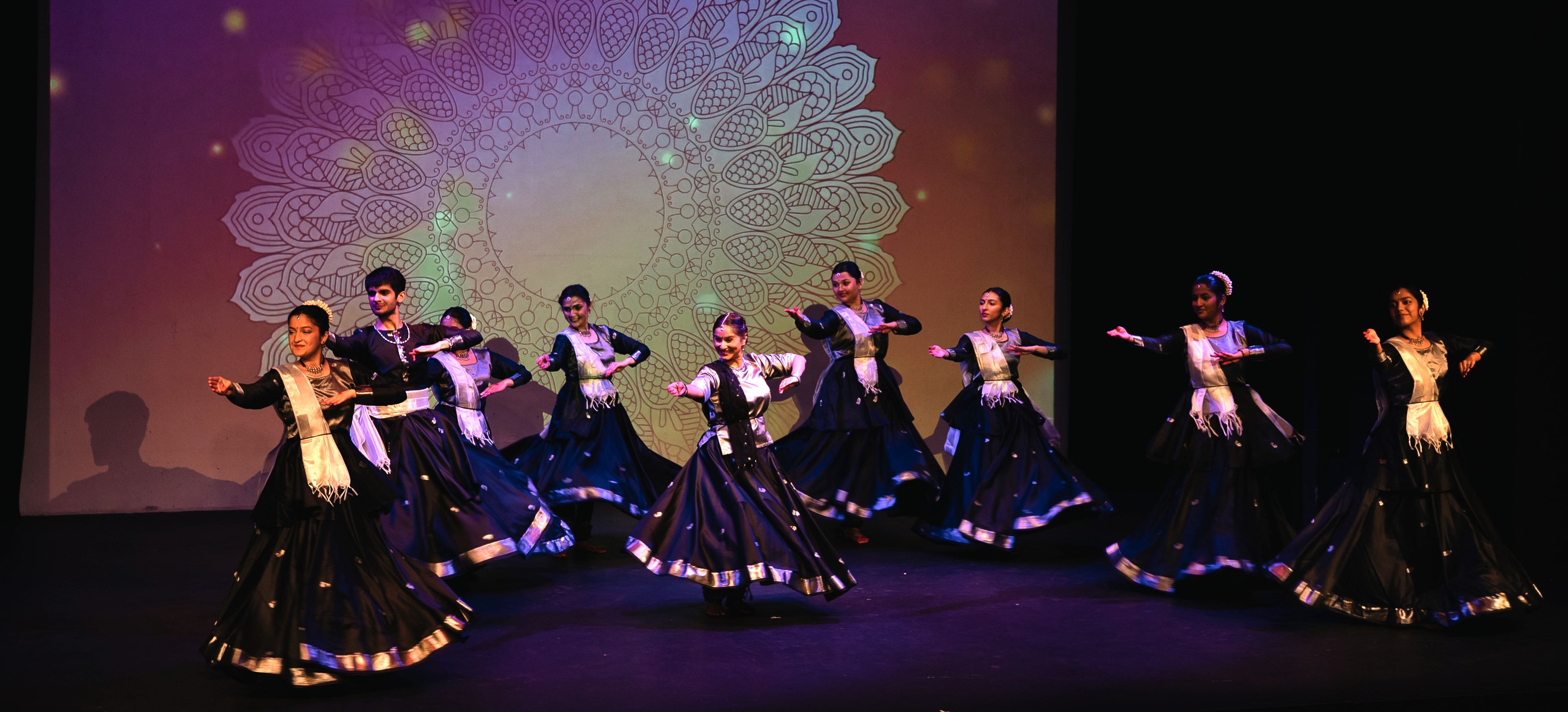 Ten Kathak dancers in dark blue and silver costumes, arms extended in unison on a darkened stage, before a large illuminated mandala projection.