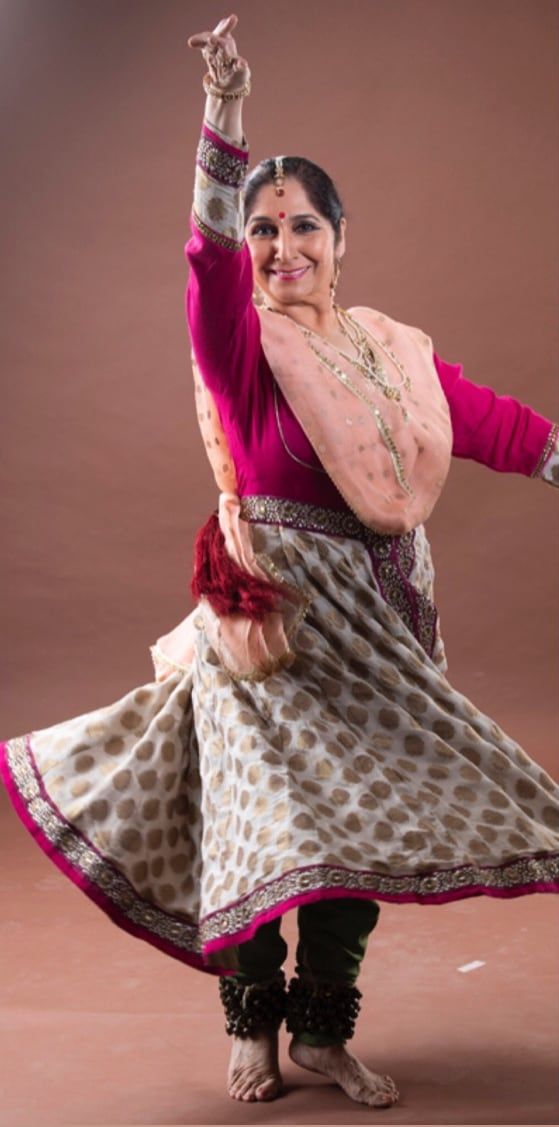 Pandita Uma Dogra ji mid-pose in a pink choli and printed anarkali, one arm raised, ghungroos at her ankles